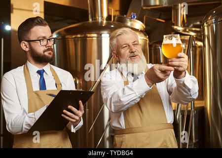 Two brewery specialists looking at beer glass, analyzing. Elderly brewer holding glass with cold beer with foam. Handsome bearded man, expert holding folder, looking at beer, writing. Stock Photo