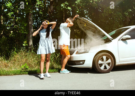 The young couple broke down the car while traveling on the way to rest. They are trying to fix the broken by their own or should hitchhike, getting nervous. Relationship, troubles on the road, vacation. Stock Photo