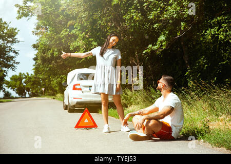 The young couple broke down the car while traveling on the way to rest. They are trying to fix the broken by their own or should hitchhike. Relationship, troubles on the road, vacation. Stock Photo