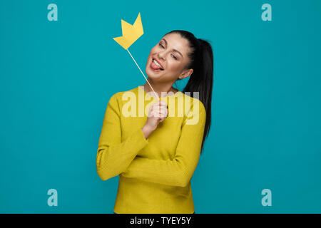 Funny young girl in yellow sweater keeping paper crown and showing tongue on blue isolated background. Excited woman looking at camera and posing in studio. Concept of happiness and fun. Stock Photo