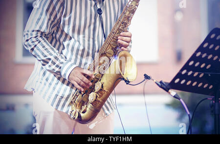 A professional musician, dressed in a striped shirt and white trousers, plays a jazz composition on a vintage gold saxophone, standing on the stage an Stock Photo