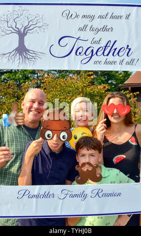 Three generations pose in a clever cut-out photo frame for a funny family portrait at an American family reunion on a summer day in Connecticut, USA. The older attendees at a typical family reunion are generally grandparents, parents, siblings and first cousins, while the youngest could be second, third or fourth cousins. Some family reunions are held on a regular annual basis, or they can be special occasions every 5th or 10th year or so. It is always a time to meet the newest members of the extended family and to share stories, photographs and food. Stock Photo