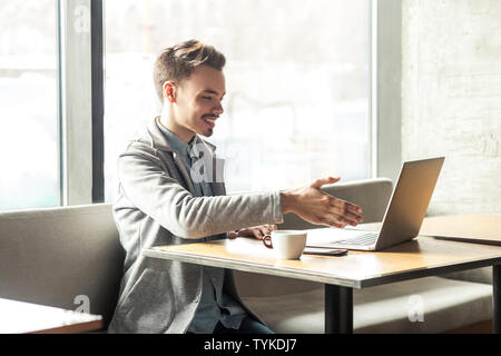 Welcome! Portrait of satisfied happy young man in grey blazer are sitting in cafe and greeting a worker through a webcam and showing handshake gesture Stock Photo