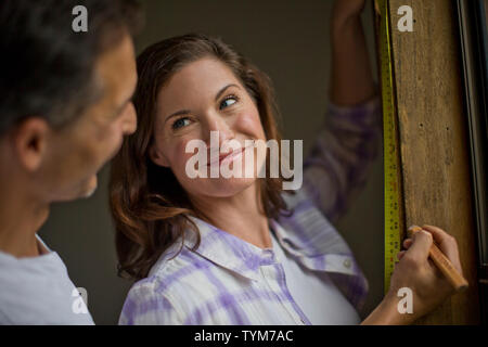 Young woman takes a break from measuring the window sill to smile lovingly at her handsome husband who has just come up behind her. Stock Photo