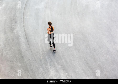 Top view of back side of young attractive athletic woman speed running on city asphalt and workout in the morning time on a sunny summer day. Female r Stock Photo