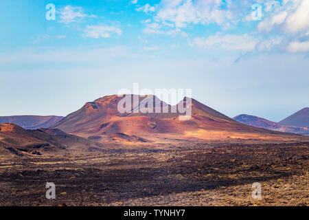 A View of the red volcanic hill in the sandy desert in Timanfaya National Park. Blue sky with clouds is in the background. It is situated in Lanzarote Stock Photo