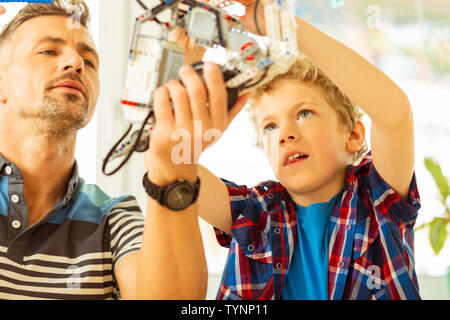 Exciting occupation. Nice pleasant father and son being busy while constructing a robot together Stock Photo