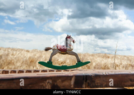 antique, rocking horse is standing on a wooden fence overlooking a field of grain Stock Photo