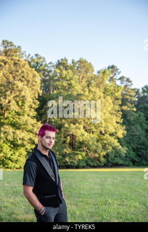 Alternative diverse male - black clothes, pink hair smirking at camera. Happy and friendly knowing smile. Punk rocker outdoors in nature - room for co Stock Photo
