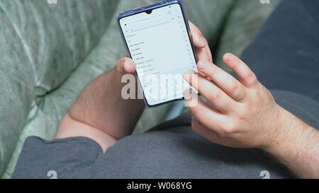 Man lying on couch and holding Huawei Mate 20 X smartphone in one hand. He is typing some text in searching line and proposed variants are seen Kyiv, Stock Photo