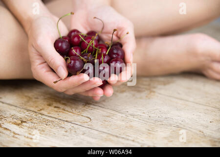 Woman sitting cross-legged on the light wooden table with her hands together holding a large handful of cherries Stock Photo