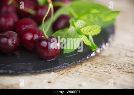 Fresh cherries on a black plate of wet slate with sprig of fresh green mint on light wooden table in selective focus for copy space Stock Photo