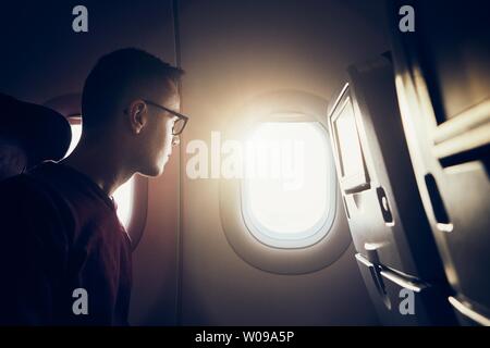 Man traveling by airplane. Pensive passenger looking through window during flight at sunset. Stock Photo