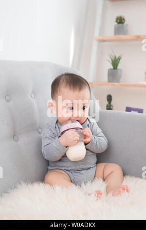 Little cute baby girl sitting in room on sofa drinking milk from bottle and smiling. Happy infant. Family people indoor Interior concepts. Childhood b Stock Photo