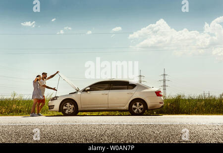 The young couple broke down the car while traveling on the way to rest. They are trying to fix the broken by their own or should hitchhike, getting nervous. Relationship, troubles on the road, vacation. Stock Photo