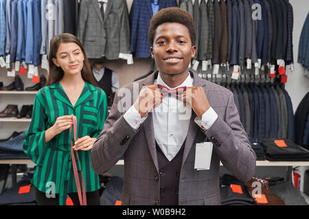 Client choosing suit, dressing white shirt, waistcoat and jacket, looking at camera and smiling. Handsome man with curly hair touching red bow tie, posing. Pretty seller holding measuring tape. Stock Photo