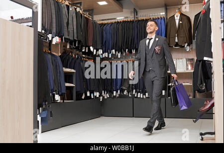 Happy client in striped suit, white shirt, necktie in boutique with shopping bags. Elegant gentleman with beard doing shopping in store. Fashionable clothing and shoes for men. Stock Photo