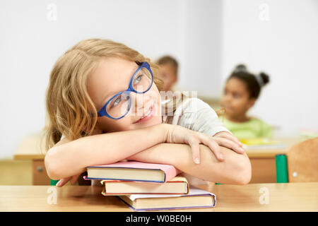 Pretty schoolgirl sitting at desk in classroom, leaning head on stack of books. Cheerful, beautiful, young pupil, teenager posing, looking away, smiling. Stock Photo