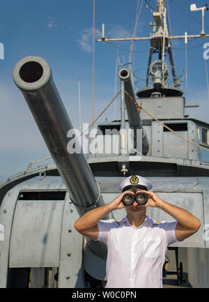 Captain of warship standing beside cannons and looking ahead. A navy officer in white uniform looking with binocular aboard a battleship. Stock Photo