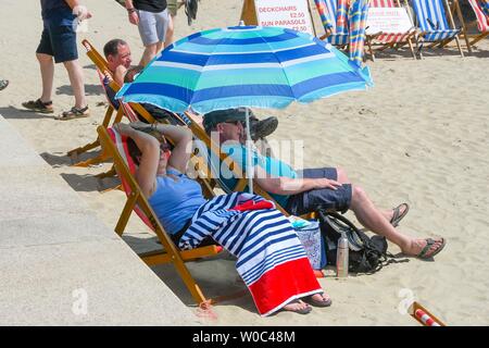 Lyme Regis, Dorset, UK.  27th June 2019. UK Weather.  Sunbathers shade themselves under an umbrella at the seaside resort of Lyme Regis in Dorset as they enjoy a day of clear blue skies and scorching sunshine.   Picture Credit: Graham Hunt/Alamy Live News Stock Photo