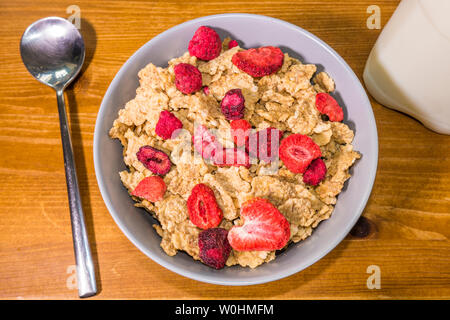 A bowl of cereal flakes mixed with raspberries and strawberries dried fruit, with a spoon and milk alongside. Stock Photo