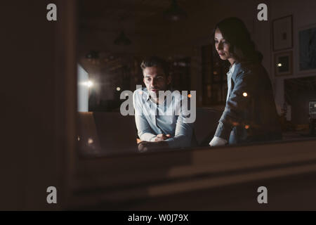 Two young work colleagues using a laptop together at a desk late at night behind office building windows reflecting the city Stock Photo