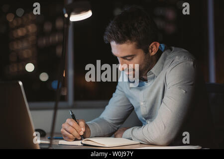 Young businessman writing notes and using a laptop while working overtime at his office desk late in the evening Stock Photo