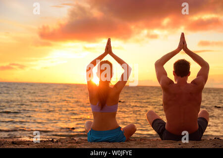 Yoga couple relaxing doing meditation on beach. Silhouettes of man and woman people practicing yoga pose sitting at a beach in the lotus position with their hands raised against a colorful sunset sky. Stock Photo