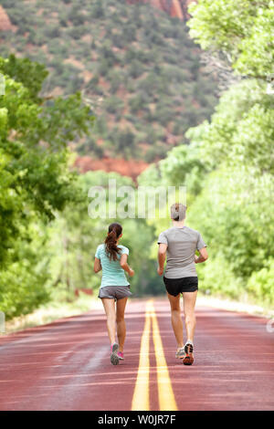 Runners running on road in nature away from camera. Couple, woman and man jogging for a run outside in amazing mountain landscape. Full body length rear view of back. Fitness and healthy lifestyle. Stock Photo
