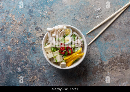 Hot noodle soup with chili, tofu, green onion and mushrooms in dish with sticks on concrete blue background. Horizontal top vie Stock Photo