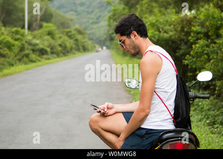 Young tourist with sunglasses sitting on a rental motorbike checking a smartphone on summer Stock Photo