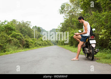 Young tourist rider sitting on a rental motorbike using a smartphone in the middle of a road Stock Photo