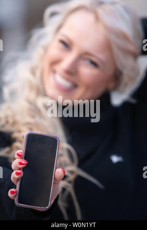 Happy laughing woman holding out her mobile phone with the blank screen to the camera and focus to her hand Stock Photo