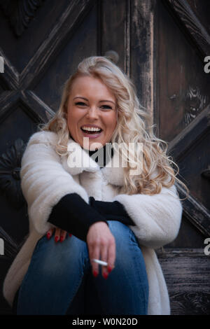 vivacious blond woman smoking outdoors sitting in front of an old wooden door laughing at the camera Stock Photo