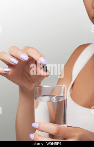 Drink medicines. Pretty young woman who takes a black pill and holds a glass of water. Isolated on a gray background. The concept of health care. Stock Photo