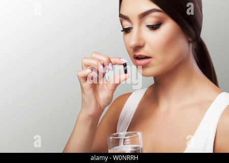 Drink medicines. Pretty young woman who takes a black pill and holds a glass of water. Isolated on a gray background. The concept of health care. Stock Photo