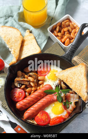 Breakfast in English scrambled eggs with bacon, beans, mushrooms, sausages, tomatoes and juice on light stone table. Stock Photo