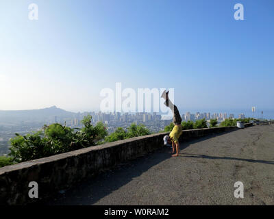 Man wearing a hat, t-shirt, pants, and slippers Handstands at Tantalus Mountain lookout point above the City of Honolulu and Diamond Head on Oahu, Haw Stock Photo