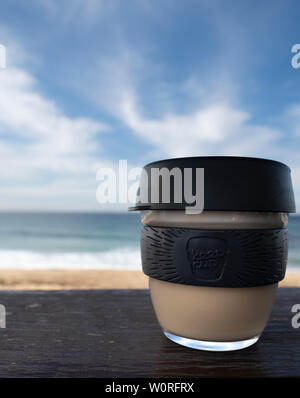 NEWCASTLE, AUSTRALIA - June 16 2019 Close up of a glass Keepcup with black trimming, filled with coffee, on a timber bench with the Pacific Ocean in t Stock Photo