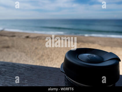 NEWCASTLE, AUSTRALIA - June 16 2019 Close up of a glass Keepcup with black trimming, filled with coffee, on a timber bench with the Pacific Ocean in t Stock Photo