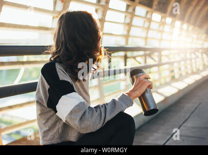 Slender young brunette woman street dancer gymnast sitting and rest with thermo cup on the street bridge at sunrise Stock Photo