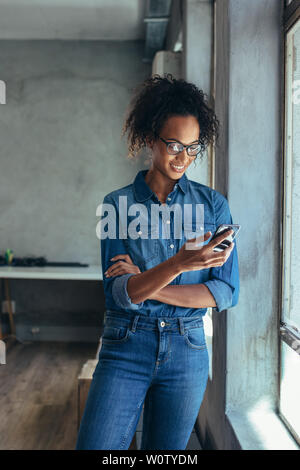 Smiling businesswoman using mobile phone standing in office. Woman entrepreneur standing by window with a cell phone. Stock Photo