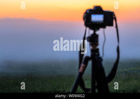 black digital camera on tripod shooting foggy morning landscape at summer with selective focus. Stock Photo