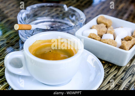 Cup of Traditional Italian Style Espresso Coffee With A Sugar Bowl on a Table Top and A Cigarette In A Glass Ashtray Stock Photo