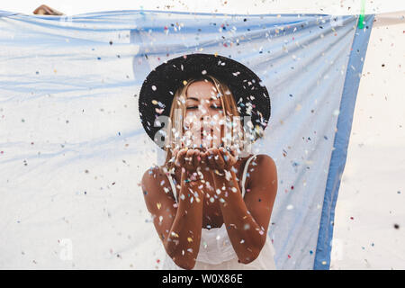 pretty woman with colorful straw hat blowing confetti in the middle of clothes hanging out to dry on a terrace in the city Stock Photo