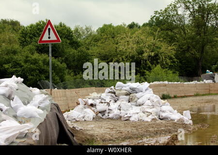 Pile of sandbags next to warning exclamation road sign and temporary flood protection wall made of box barriers covered with thick geotextile fabric Stock Photo