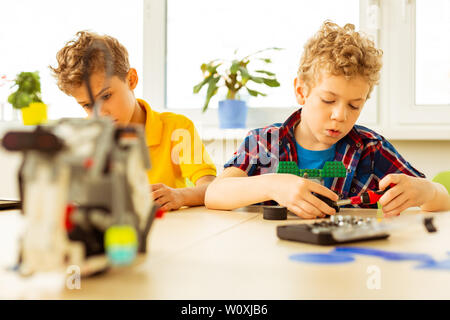 Two classmates. Nice blonde boy sitting with his friend while constructing a toy Stock Photo