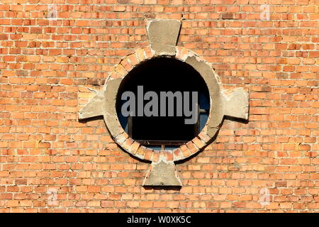 Round window opening with partially broken glass and decorative red bricks tiles frame on old abandoned industrial complex building Stock Photo