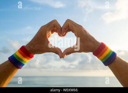 Hands wearing LGBTQI pride rainbow colors sport wristbands making a heart gesture above a golden sunset sea horizon Stock Photo