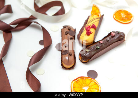 A close-up of a set of three eclairs with different fillings and design isolate on a white surface decorated with chocolate drops, dried oranges and b Stock Photo